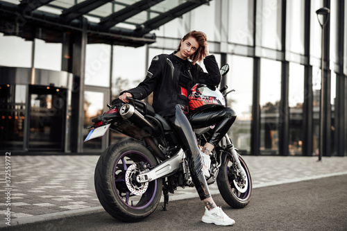 Beautiful young brunette lady in a black motorcycle jacket sits on a purple sport motorbike