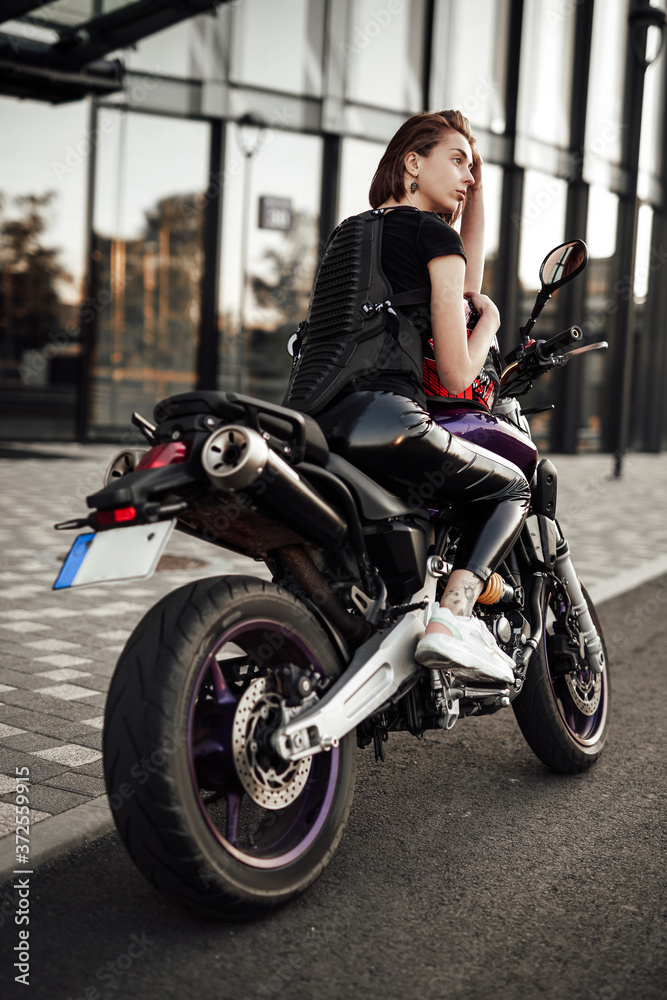 Beautiful young brunette girl in a black motorcycle jacket sits on a purple sport motorbike