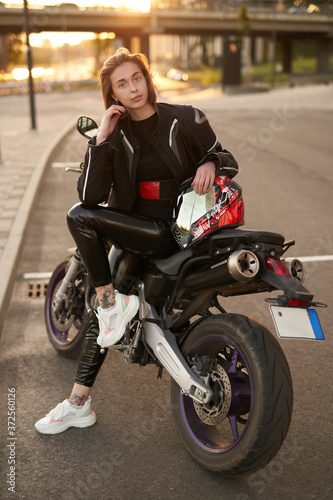 Young brunette girl in a black motorcycle jacket sits on a purple sport motorbike, with the sunset on the background © Fxquadro