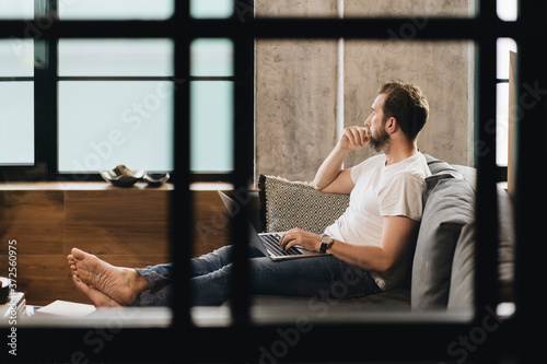 Matur man sitting on couch with laptop on lap, looking away, thinking photo