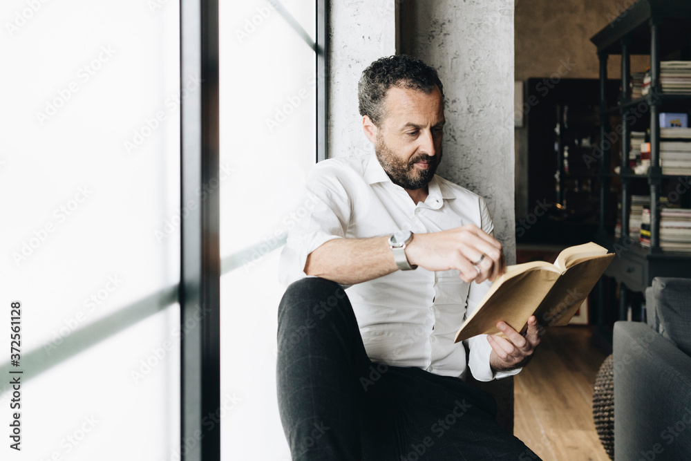 Mature man sitting on window sill, reading book Mature man sitting on window sill, reading book