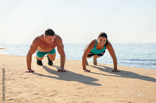 A couple doing exercise on the beach near the sea, doing plank smiling at each other. Sport in the nature to stay healthy and fit. Sunny beach day workout.