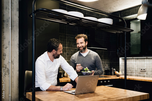 Mature friends working on laptop in kitchen, one eating salad
