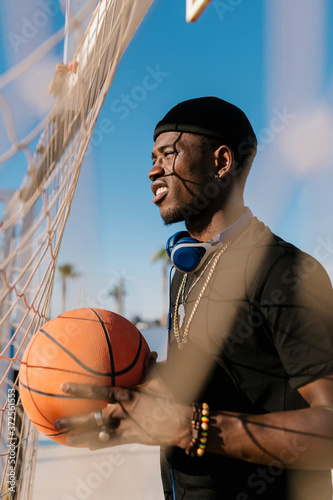 Man holding basketball looking away while standing by net in court during sunny day photo