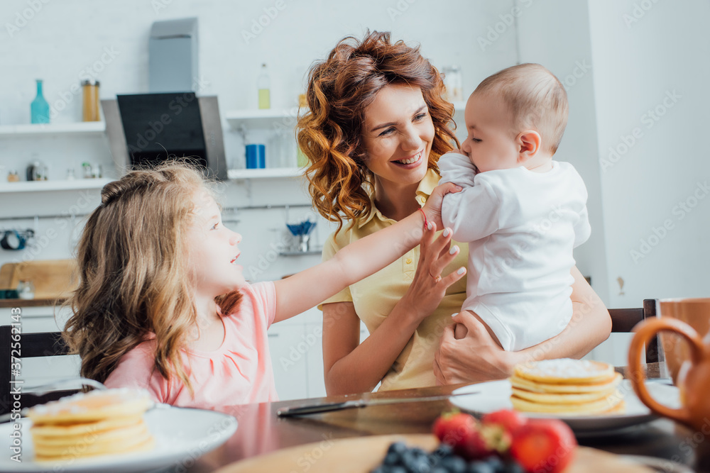 selective focus of young mother with infant boy and daughter sitting at table served with breakfast