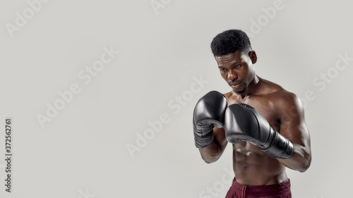 Young muscular african american male boxer looking down, wearing boxing gloves, standing isolated over grey background. Sports, workout, bodybuilding concept © Svitlana