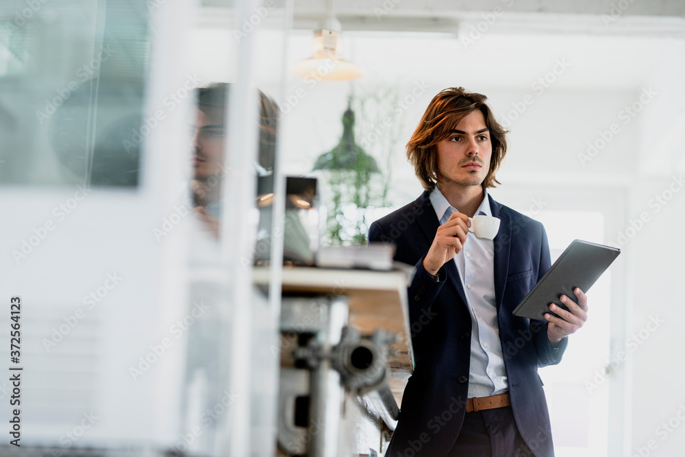 Thoughtful male entrepreneur holding digital tablet and coffee cup while standing in cafe Thoughtful male entrepreneur holding digital tablet and coffee cup while standing in cafe