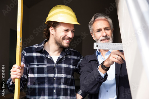 Smiling construction worker looking at measurement showing by architect in constructing house seen through window