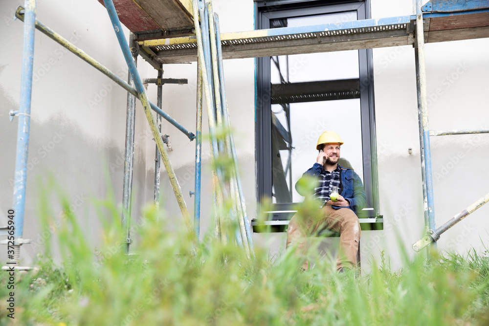 Construction worker talking over smart phone while sitting against window at site Construction worker talking over smart phone while sitting against window at site