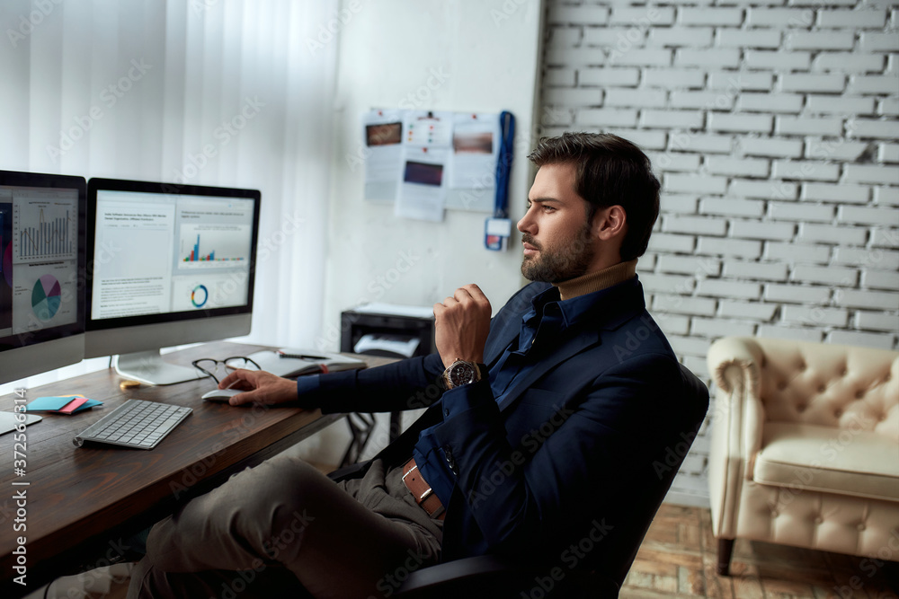 Side view of young successful businessman or sales manager analyzing financial data on computer, looking at statistics on pc screen while sitting at his workplace in the modern office