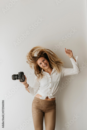 Cheerful woman holding camera shaking head while standing against white wall photo