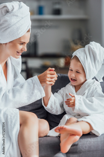 selective focus of young mother making manicure to child while sitting together in white bathrobes and towels on heads
