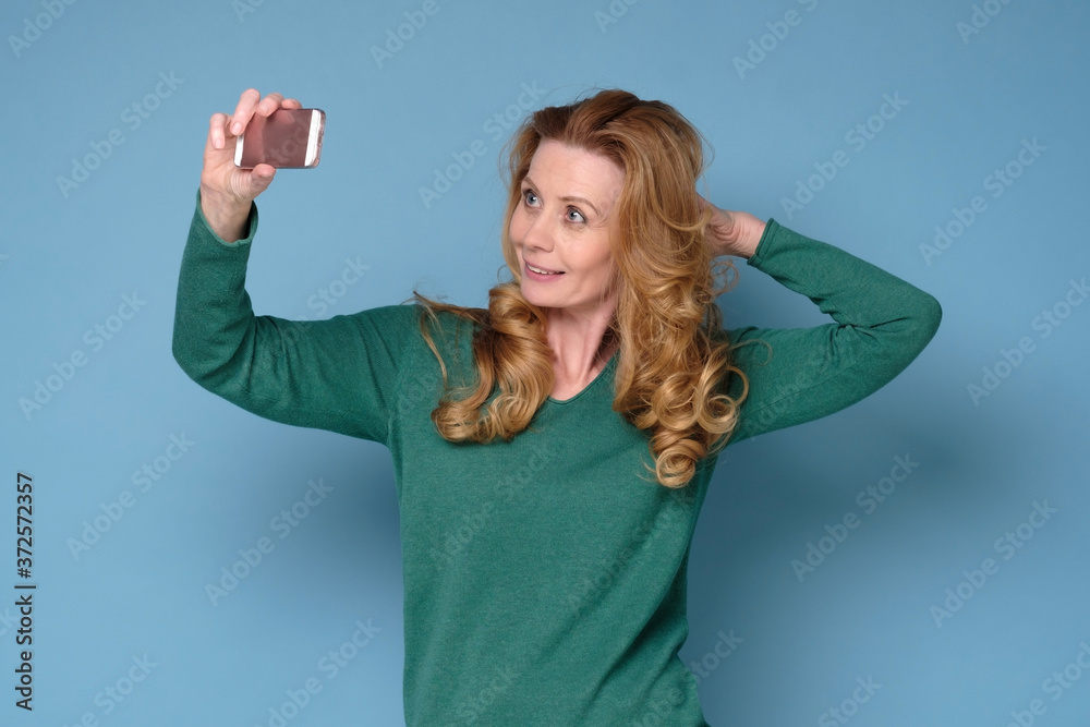 Obraz premium Caucasian mature woman take a self portrait with her smart phone. Studio shot on blue wall.