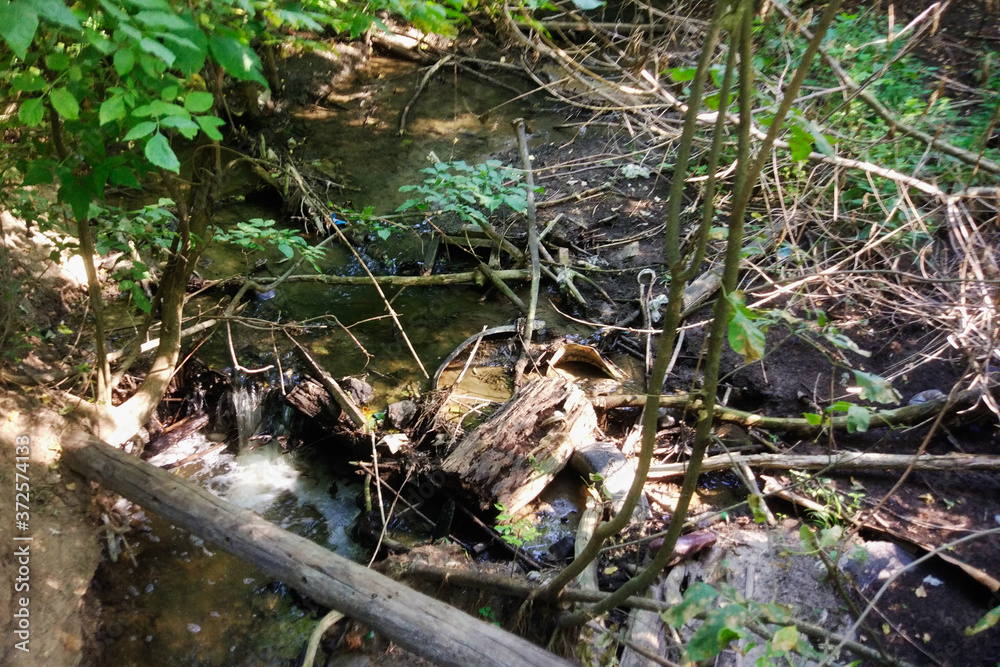 A small forest river is polluted with garbage. Plastic trash, foam, wood and dirty waste