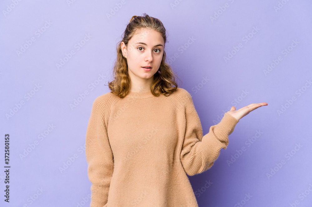 Young caucasian woman on purple background impressed holding copy space on palm. Young caucasian woman on purple background impressed holding copy space on palm.