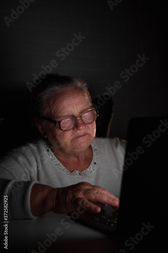 Senior Woman Using Laptop In Dark room. Elderly woman's hands on a computer keyboard in the dark, light from the screen. The older generation is searching for information and working 