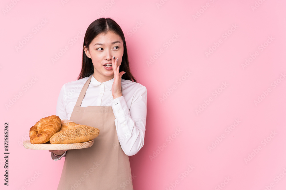 Young chinese baker woman isolated is saying a secret hot braking news and looking aside