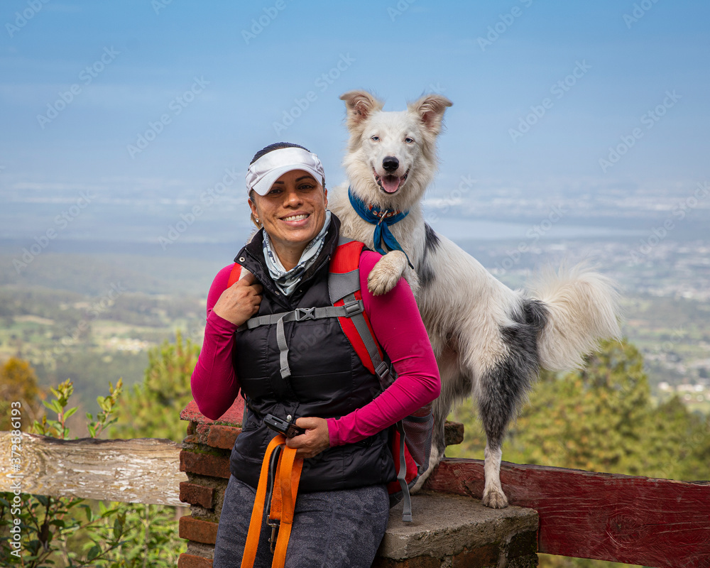 smiling mexican woman with happy white border collie dog standing behind with paws on her shoulder, landscape behind them