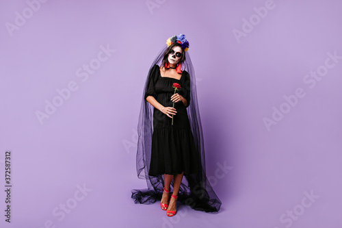 Mexican widow sadly looks into camera, holding red rose. Full-length photo of woman in black outfit with bridal veil