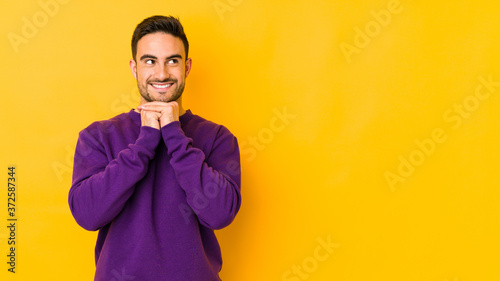 Photography Young caucasian man isolated on yellow bakground keeps hands under chin, is looking happily aside