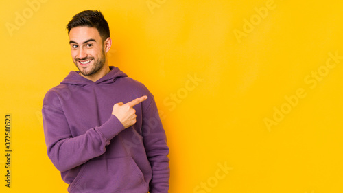 Obraz na płótnie Young caucasian man isolated on yellow bakground smiling and pointing aside, showing something at blank space