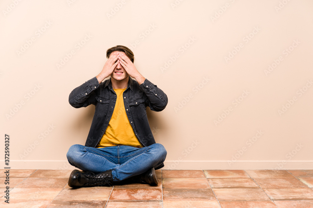 Young caucasian man sitting on the floor isolated covers eyes with hands, smiles broadly waiting for a surprise. Young caucasian man sitting on the floor isolated covers eyes with hands, smiles broadly waiting for a surprise.