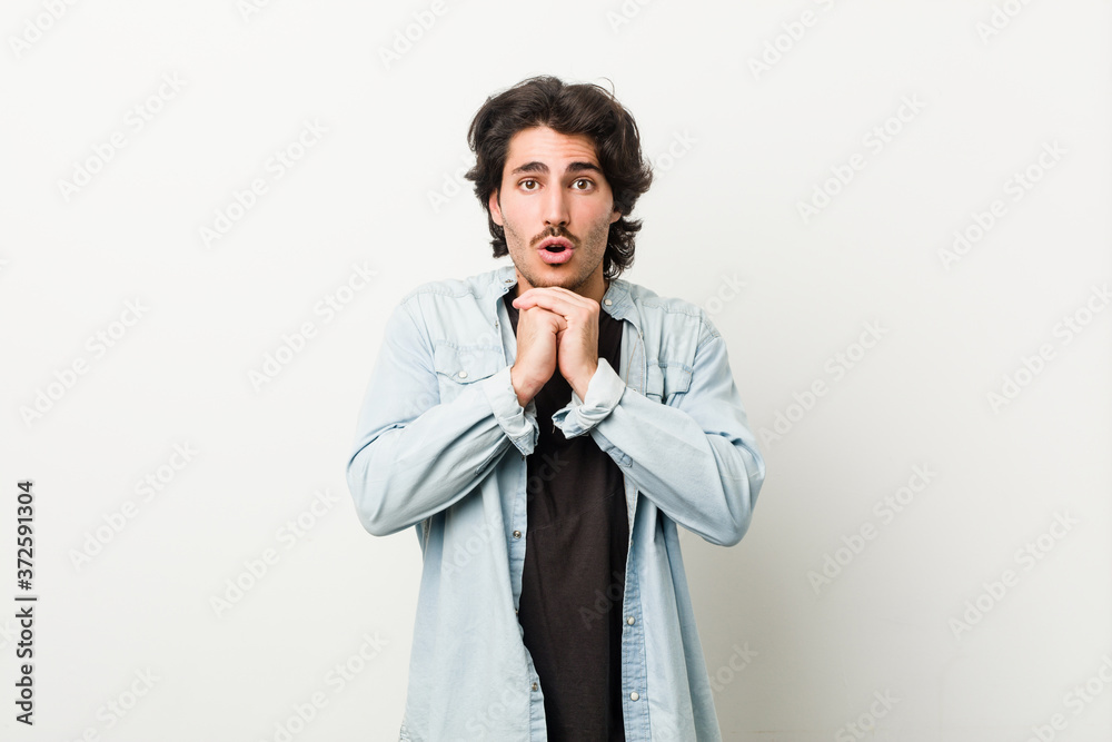 Young handsome man against a white background praying for luck, amazed and opening mouth looking to front.