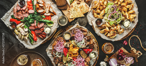 Assorted snacks for beer in plates, delicious grilled meat, vegetables, fries potatoes, smoked sausages, bbq sauce, chips, bread, pita and beer in glass on dark background, top view, toning