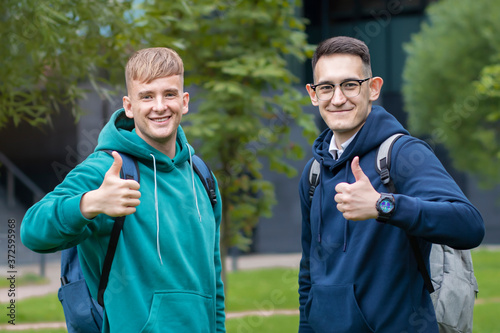 Two 2 handsome happy young men different nationalities outdoor. University or college multinational students with backpacks showing thumb up, like approval gesture. Boys are smiling, looking at camera