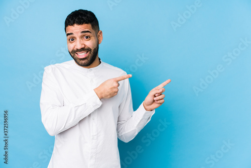 Young latin man against a blue background isolated shocked pointing with index fingers to a copy space.