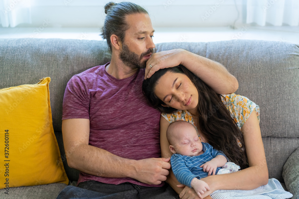 Fototapeta premium Beautiful Caucasian mother holding newborn baby son and sleeping together on sofa. Man kissing and embracing his wife and cute infant child boy with love. Happy Family and baby healthcare concept.