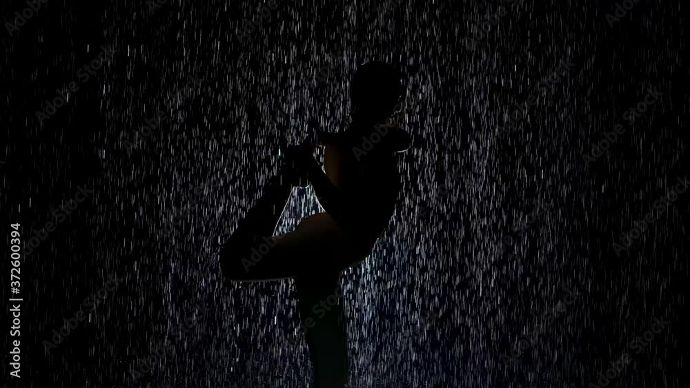 Dancer pose in the pouring rain. Wet body silhouette of young woman practicing yoga exercises in dark studio with neon refreshing white. Slow motion. Dancer pose in the pouring rain. Wet body silhouette of young woman practicing yoga exercises in dark studio with neon refreshing white. Slow motion.