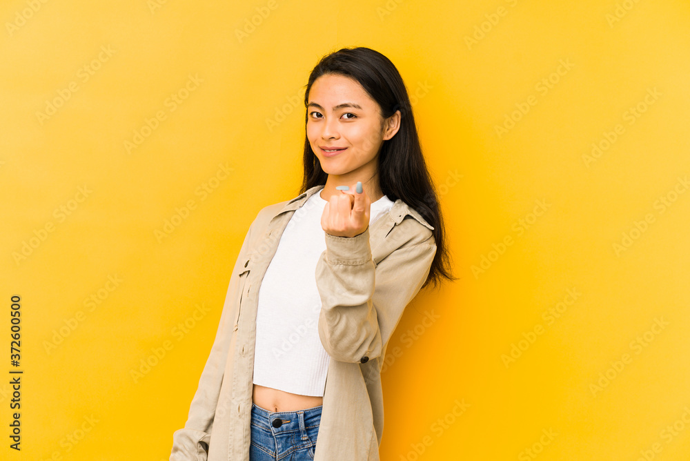 Young chinese woman isolated on a yellow background pointing with finger at you as if inviting come closer. Young chinese woman isolated on a yellow background pointing with finger at you as if inviting come closer.