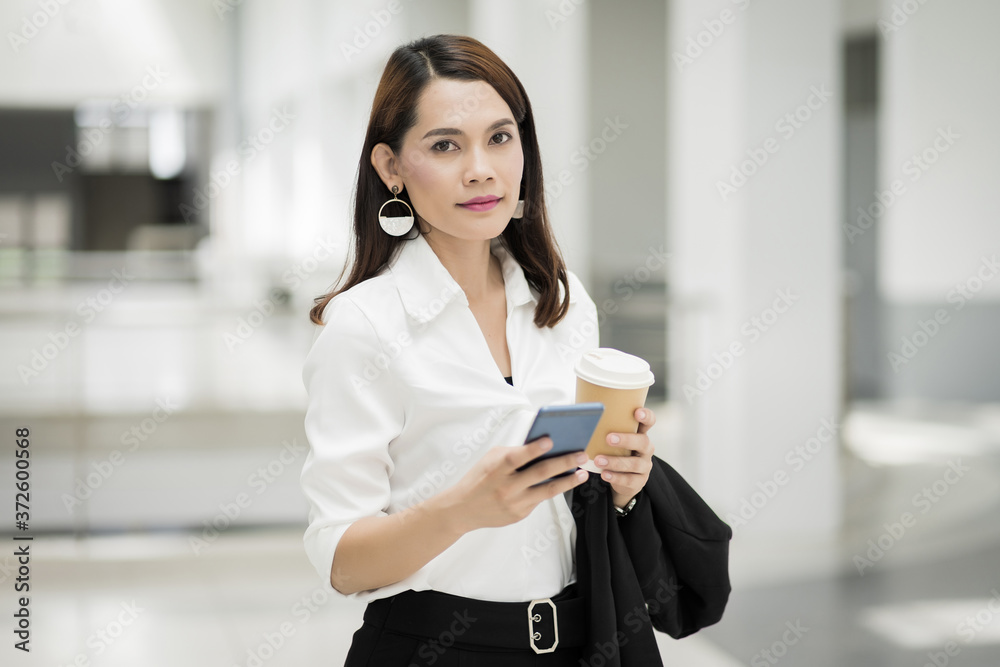 Portrait of a confident businesswoman in business suit holding a cup of coffee while using cellphone during break-time in the business building. Business stock photo. Portrait of a confident businesswoman in business suit holding a cup of coffee while using cellphone during break-time in the business building. Business stock photo.