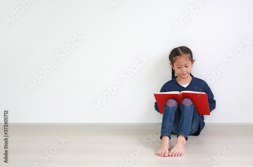 Portrait asian little kid girl in school uniform reading a book and sitting against white wall in the room with copy space.