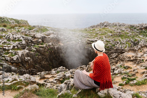 Back view of unrecognizable female traveler in stylish knitwear and hat meditating with mudra gesture while sitting on stone in front of Jesters of Arenillas geyser during trip to Asturian coast of Spain photo