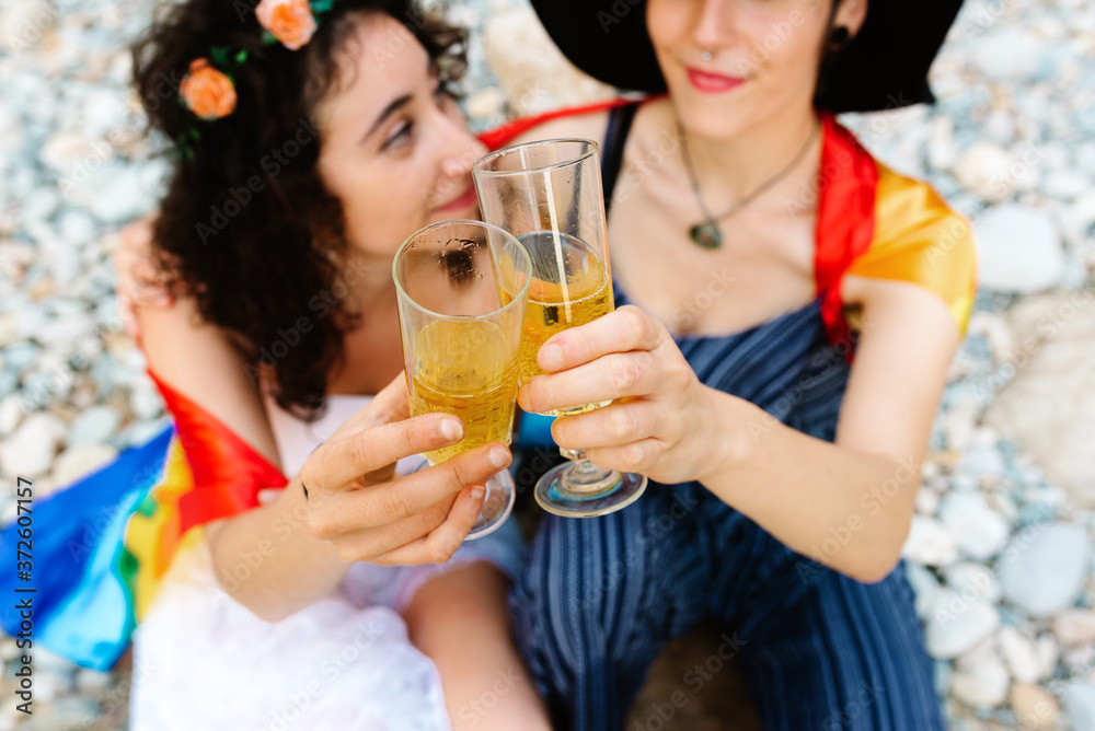 High angle of tender lesbian couple sitting on stony beach and covering with rainbow flag while embracing and clinking glasses with champagne High angle of tender lesbian couple sitting on stony beach and covering with rainbow flag while embracing and clinking glasses with champagne