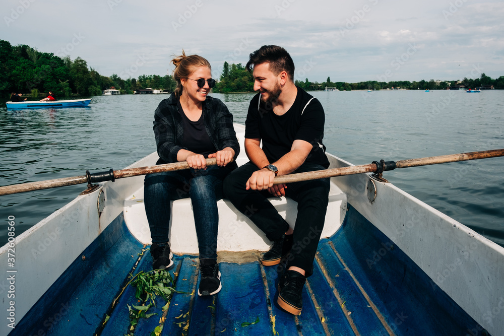 Full body happy smiling couple in black casual outfits rowing boat with oars on calm beautiful pond while looking at each other during clear summer day in Saint George, Transylvania, Romania Full body happy smiling couple in black casual outfits rowing boat with oars on calm beautiful pond while looking at each other during clear summer day in Saint George, Transylvania, Romania