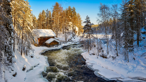Small cozy wooden house located in picturesque forest covered by snow near wild river against cloudless blue sky