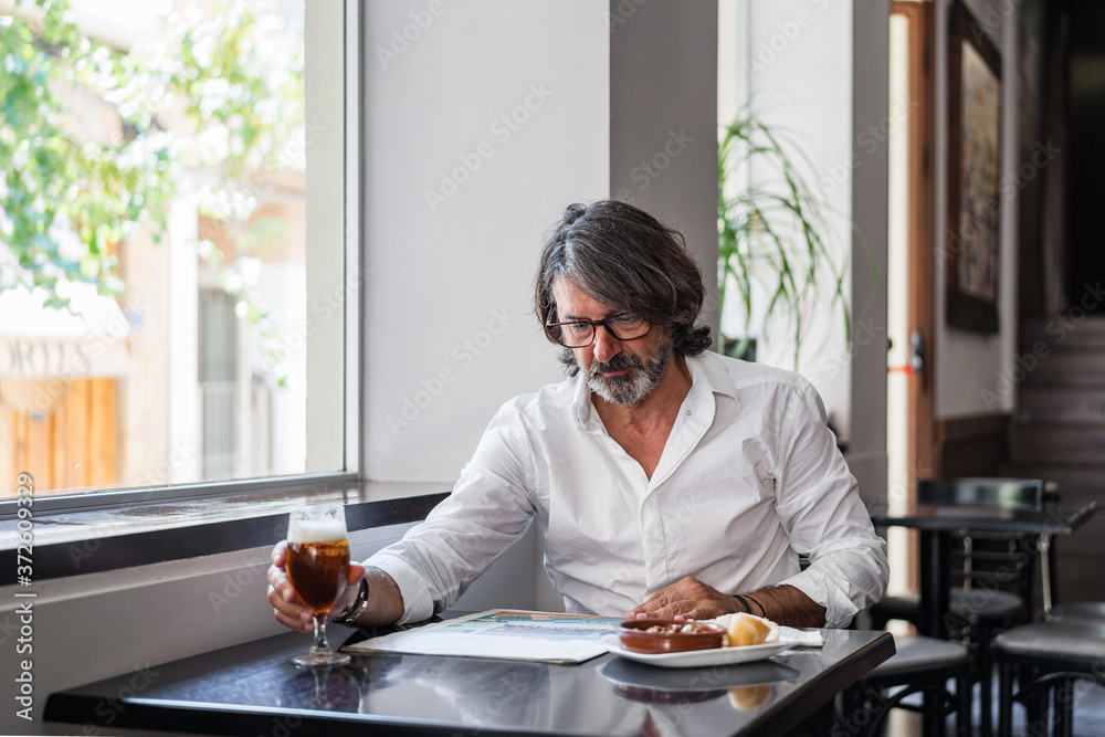 Positive ethnic bearded aged man sitting at table with of glass of foamy beer and reading newspaper near window Positive ethnic bearded aged man sitting at table with of glass of foamy beer and reading newspaper near window