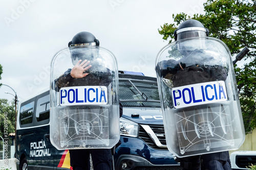 Low angle of anonymous police soldiers in protective uniforms and helmets standing against squad van and defending by riot shields