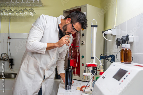 Adult male scientist in uniform and eyewear measuring sugar content of wine while using transparent wine meter in laboratory standing near electronic equipment