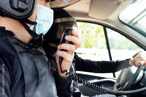 Crop unrecognizable policeman in medical mask and protective gear talking on radio set while sitting in car with partner
