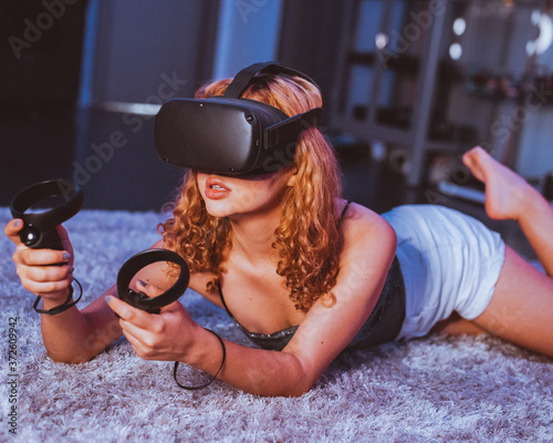 Seductive barefoot anonymous female in casual wear lying with open mouth on soft plaid on bed while playing game with controllers and in goggles of virtual reality photo