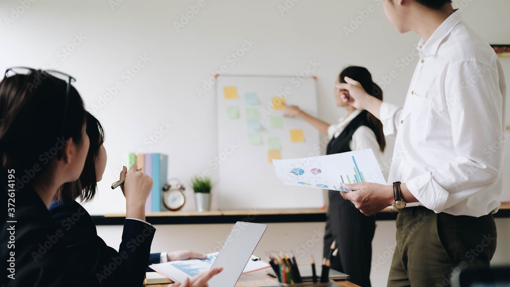 Naklejka premium Businesswoman sticking adhesive notes on whiteboard while colleagues standing with arms crossed at office