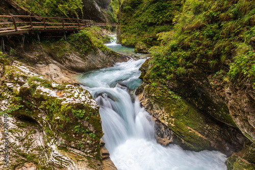Wide angle view of the Slovenian gorge of Vintgar, with a torrent running between rocks and into rapids surrounded by vegetation, with a wooden walkway on the left © Roberto