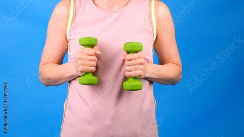 Female in a pink sports suit with dumbbells in hands does exercises on a blue background. The concept of sports, diet and weight loss
