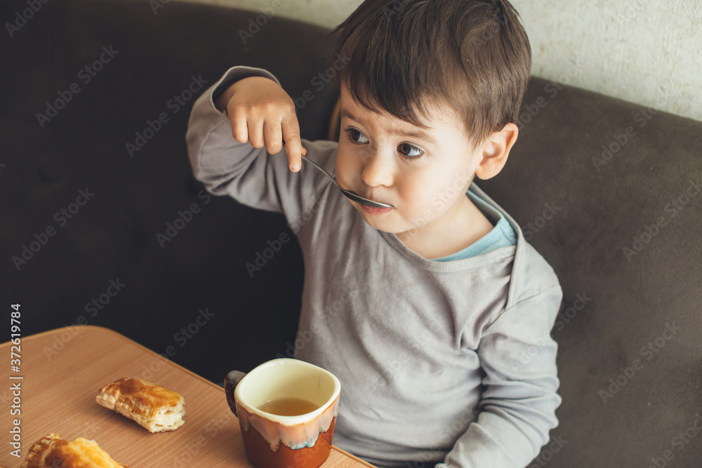 Upper photo of small caucasian brunette boy drinking a tea with spoon at the table and eating some cookies Upper photo of small caucasian brunette boy drinking a tea with spoon at the table and eating some cookies