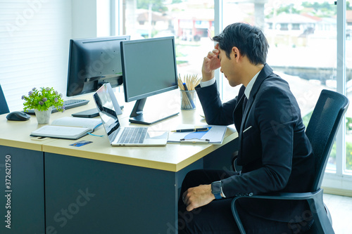 young Asian businessman in black suit sitting at the table looking a paper documents  and thinking with headache at office. Asian man working hard and feeling Stressed, side view © Nipapun