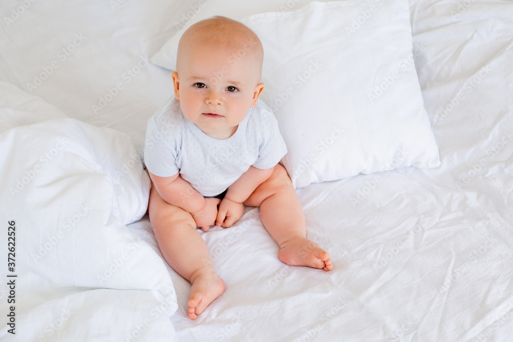 Cute baby boy 6 months smiling in a white bodysuit sitting on a bed on white bedding Cute baby boy 6 months smiling in a white bodysuit sitting on a bed on white bedding